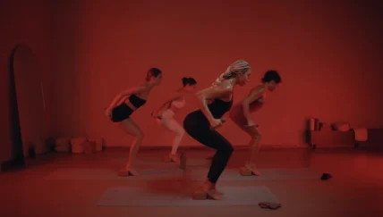 Four women perform a synchronized strength workout using dumbbells and yoga blocks in a red-lit studio.